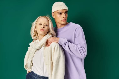 Şapkalı ve kışlık giysili genç adam turkuaz arka planda sarışın bir kadının omuzlarına dokunuyor.