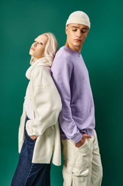 Kış kıyafetli sarışın kadın sırt sırta uzun boylu bir adamla turkuaz arka planda duruyor.