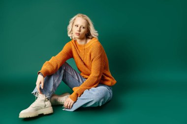 Dövmeli, hardal sarısı süveter ve turkuaz arka planda oturan şık kot pantolonlu kadın.