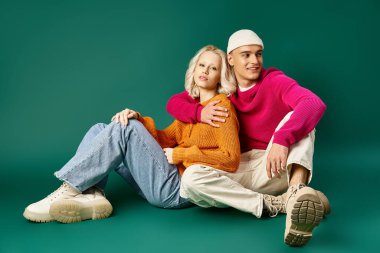 Sıcak kazaklı bir çift, bereli bir adam turkuazda birlikte otururken sarışın bir kadına sarılıyor.