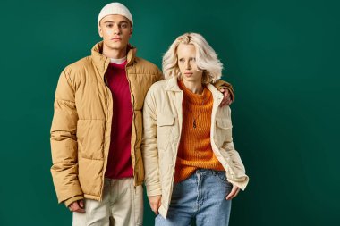 Kışlık kabarık ceketli şık bir adam kadına sarılıyor ve turkuaz arka planda birlikte poz veriyor.