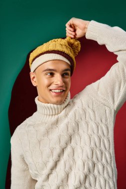 joyful man in white sweater wearing yellow mustard bobble hat on turquoise background, winter trends clipart