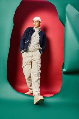 Kış kıyafetli şık bir adam elleri ceplerinde turkuaz arka planda yırtık kırmızı renkte yürüyor.