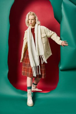 full length of blonde woman in trendy winter attire ripping turquoise paper and walking red backdrop clipart