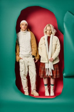 Yırtık turkuaz ve kırmızı arka planda, şık bir kadının yanında el ele poz veren genç adam.