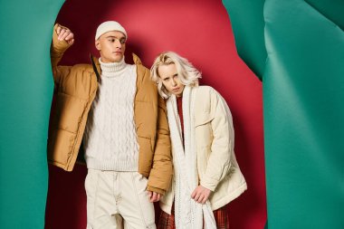 Bereli ve kışlık ceketli genç adam yırtık turkuaz ve kırmızı arka planda şık bir kadının yanında.