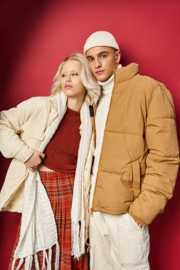 stylish man and woman in warm winter outerwear posing together on red background, winter fashion clipart