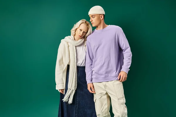 handsome tall man in beanie hat posing with blonde woman in winter attire on turquoise backdrop