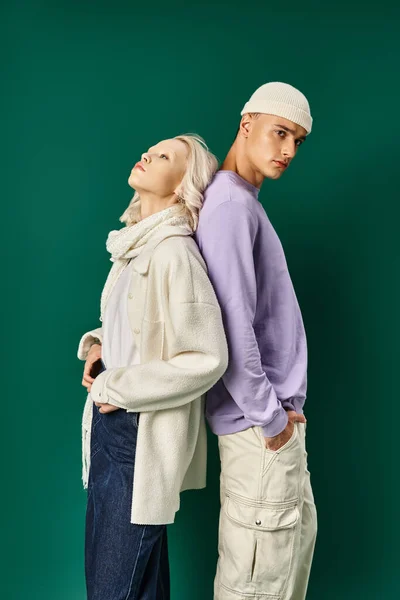 tall man in hat and winter attire standing back to back with blonde woman on turquoise backdrop