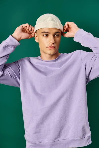 ターコイズの背景 冬のファッションにビーニーの帽子を調整する紫色のスエットシャツの良い見た目の人 — ストック写真