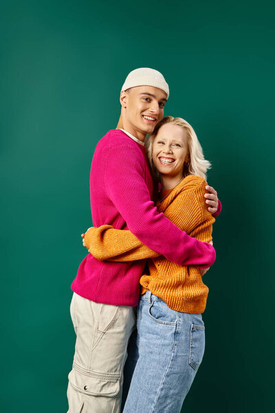 joyful couple in winter attire, man in beanie hugging blonde girlfriend on turquoise, winter fashion