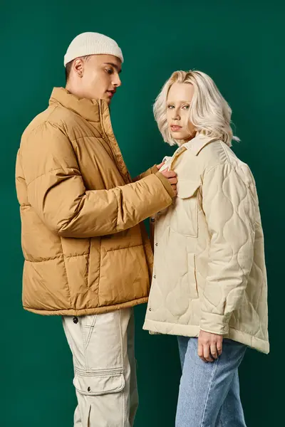 ターコイズの背景 冬のファッションでブロンドの若い女性にパッファージジャケットを着用する世話をする男 — ストック写真