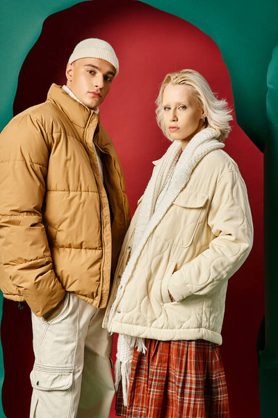 young couple in trendy winter jackets posing with hands in pockets on red with turquoise backdrop