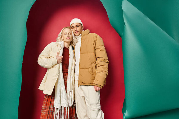 stylish young couple in warm winter outerwear posing together near torn turquoise and red backdrop