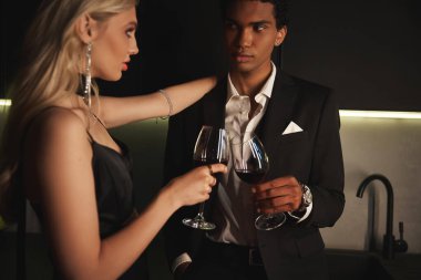 Akşam kıyafetleri içinde şarap bardakları tutan ve birbirlerine bakan çok kültürlü bir çift.
