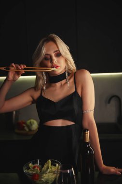 Aksesuarlı, akşam giysili çekici bir kadın mutfaktayken yemek çubukları kullanıyor.