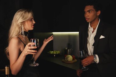 good looking multicultural couple in chic clothes having argument and holding red wine in glasses clipart