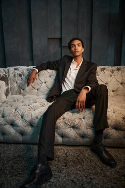 handsome young african american man in elegant suit sitting on comfy sofa and looking at camera clipart