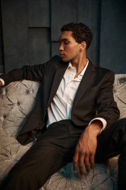 handsome young appealing african american man in black suit sitting on sofa and looking away clipart