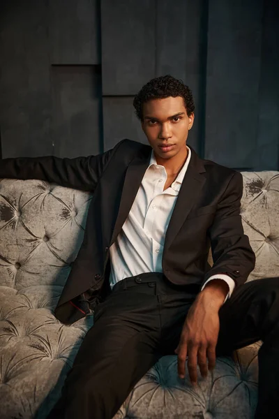 stock image handsome relaxed african american male model in chic suit sitting on sofa and looking at camera