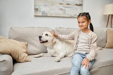 Mutlu kız koltukta oturuyor ve modern oturma odasında sevimli Labrador 'a sarılıyor, kürklü arkadaş ve çocuk