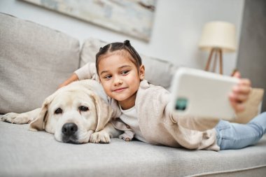 Güzel ilkokul kızı labrador köpeğiyle selfie çekiyor modern apartmanda, evcil hayvan.