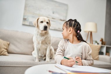 Sıradan kıyafetler içindeki küçük kız renkli kalemle çiziyor ve oturma odasında labrador arıyor, sanat eseri.