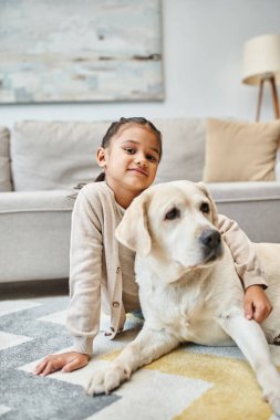 Güzel kız halının üzerinde oturuyor ve modern oturma odasında Labrador 'u okşuyor, çocuk ve tüylü arkadaşı