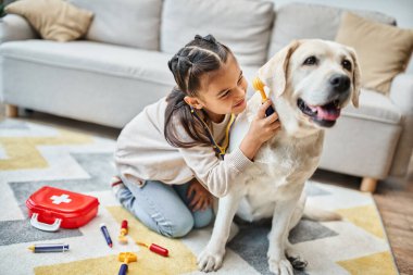 Modern oturma odasında labrador 'la doktorculuk oynayan gündelik kıyafetli tatlı kız, oyuncak ilk yardım çantası.