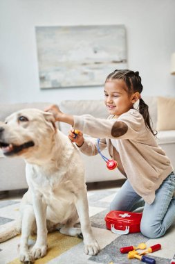 Modern oturma odasında labradorla doktorculuk oynayan sıradan giyinmiş mutlu çocuk, oyuncak steteskop.