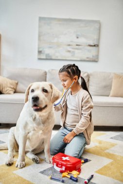 Günlük kıyafetli mutlu kız oturma odasında labrador köpeğiyle doktorculuk oynuyor, ilk yardım çantasını oyuncak ediyor.