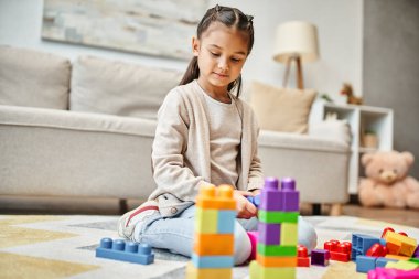 cute girl playing with colorful toy blocks on carpet in living room, building tower game clipart