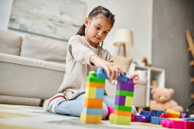 Modern oturma odasında renkli oyuncak bloklarıyla oynayan sevimli kız kule oyunu yapıyor.