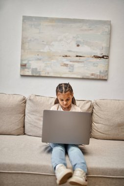 happy elementary age girl sitting on sofa and using laptop in modern living room, e-learning clipart