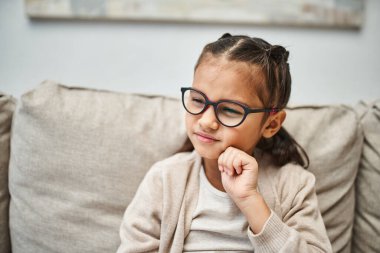pensive elementary age girl in casual attire wearing eyeglasses and looking away in living room clipart