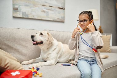 Günlük giysili ve gözlüklü mutlu çocuk oturma odasındaki kanepede labrador köpeğiyle doktorculuk oynuyor.