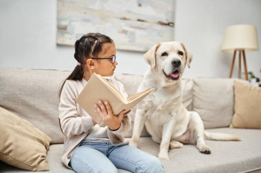 Gündelik giyinen ve gözlüklü akıllı kız oturma odasındaki kanepede Labrador Köpeğine kitap okuyor.