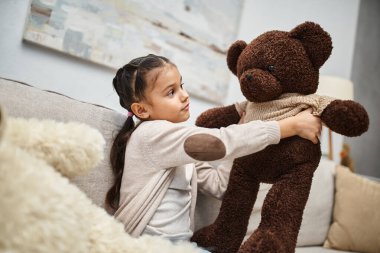 İlkokul çağındaki tatlı çocuk, gündelik kıyafetleriyle kanepede, yumuşak oyuncak ayılarla oturuyor.