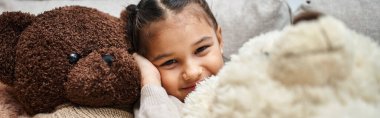 cheerful elementary age girl sitting among soft teddy bears on couch in modern living room, banner clipart