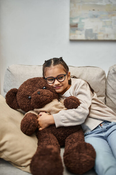 happy kid in casual wear and eyeglasses hugging soft teddy bear and sitting on sofa in living room