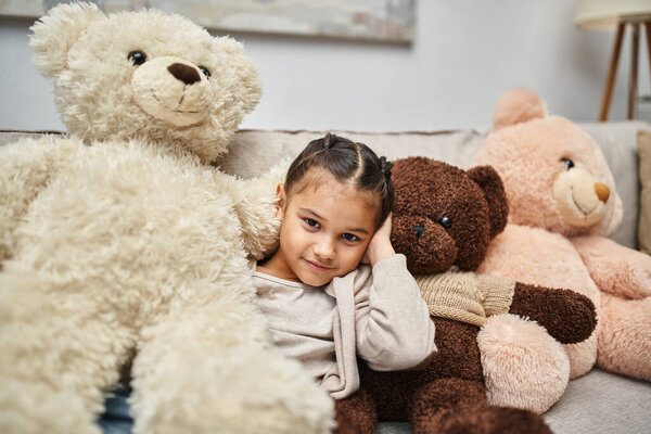 adorable elementary age girl sitting among soft teddy bears on couch in modern living room