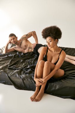 Bulanık erkek arkadaşının yanında oturan seksi iç çamaşırlı Afrikalı Amerikalı kadına odaklan.