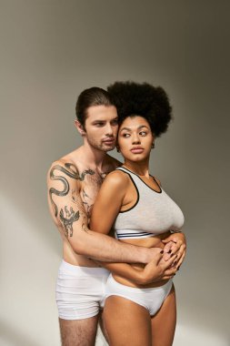 Dövmeleri olan çekici bir adam, güzel Afrikalı Amerikalı sevgilisine sarılıyor gri arka planda.