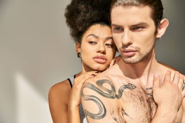 Dövmeli seksi genç adam güzel Afrikalı Amerikalı kız arkadaşına sarılıyor gri arka planda.