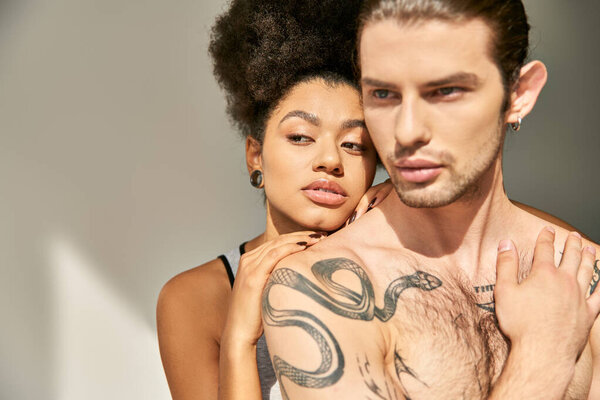 sexy young man with tattoos hugging his beautiful african american girlfriend on gray backdrop