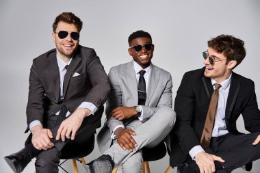 joyous young multicultural men in business smart suits sitting on chairs and smiling happily clipart