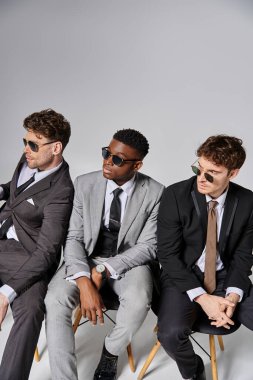handsome multiracial men with sunglasses in business attires sitting on chairs on gray backdrop clipart