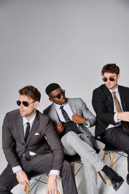 good looking diverse men with sunglasses in smart attires sitting on chairs on gray backdrop clipart