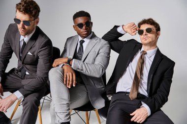 handsome interracial men with sunglasses in smart attires sitting on chairs on gray backdrop clipart