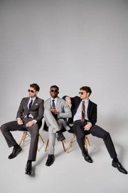 attractive interracial men with sunglasses in smart attires sitting on chairs on gray backdrop clipart